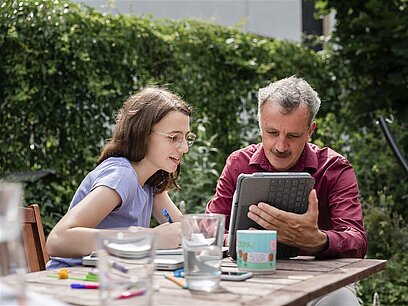 Ein Vater und seine Tochter sitzen an einem Tisch und blicken auf ein Tablet.