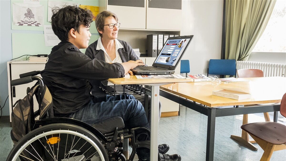 Ein Jugendlicher im Rollstuhl und eine ältere Frau sehen sich etwas auf einem laptop an.