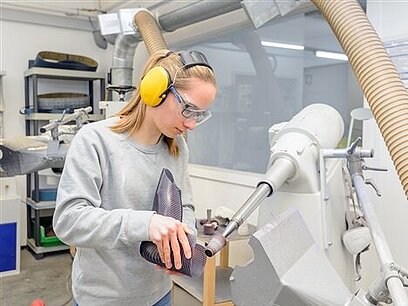 Eine junge Frau mit Gehörschutz bearbeitet ein Schuhmodell mit einer Maschine.